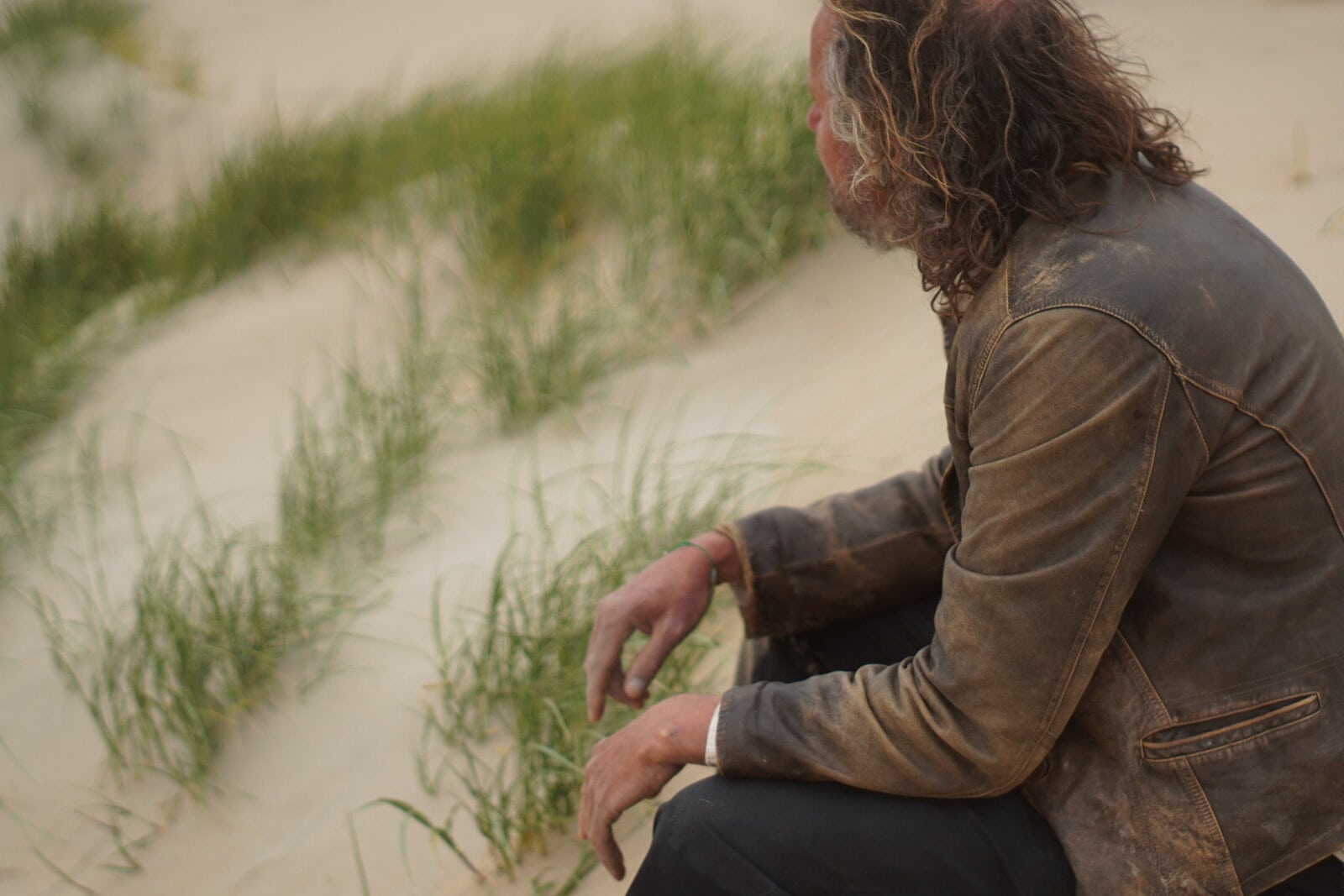 Man met lang haar en leren jas zit op het strand.